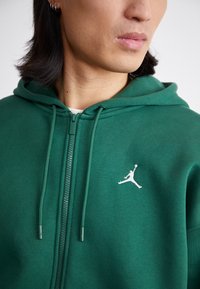 Grøn zip-up hoodie med snører, blød tekstur, hvidt logo på brystet, ribbede manchetter og søm, der tilbyder afslappet og komfortabelt brug.