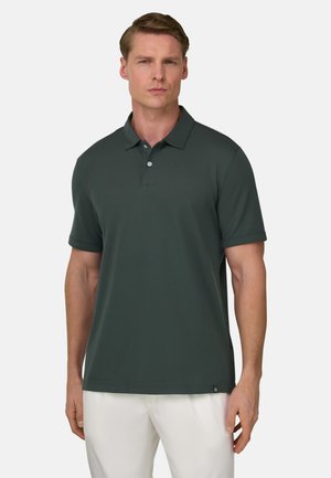 Man die een donkergroen poloshirt met korte mouwen en een witte broek draagt, staand tegen een effen achtergrond.