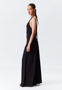 Crna halter-kragna maxi haljina s uskim korzetom, otvorenim leđima i lepršavom suknjom. Glatka, lagana tkanina prikazuje elegantnu siluetu.