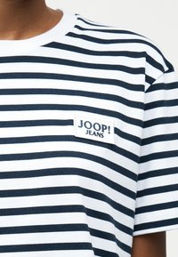 Λευκή και ναυτικό μπλε ριγέ οριζόντια μπλούζα με ετικέτα "JOOP! JEANS" στο στήθος, φορεμένη από άτομο με μέτριο-σκούρο τόνο δέρματος.