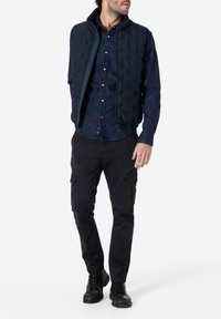 Marineblauer gesteppter Weste über einem dunklen Jeanshemd, kombiniert mit dunklen Cargo-Hosen und schwarzen Stiefeln. Verfügt über Reißverschlüsse und Taschen für praktische Nutzung.