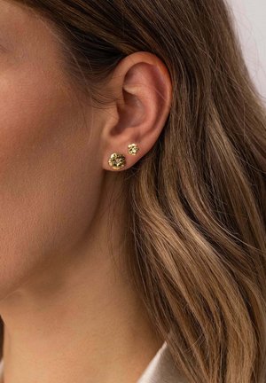 Nahaufnahme eines Ohrs mit zwei goldfarbenen, blumenförmigen Steckerohrringen, getragen in hellbraunem, welligem Haar.