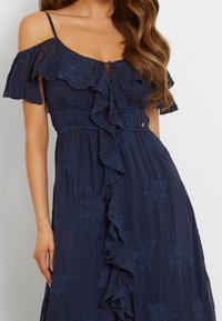 Navy blue dress with off-shoulder ruffle sleeves, featuring floral embroidery, a ruffled front, and a fitted waist.