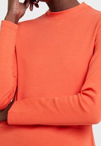 Långärmad topp i ljus korall, tillverkad av mjuk bomullsmix. Har en hög halsringning och åtsittande ärmar, med en slät textur och inga mönster.