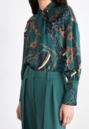 Kobieta w zielonej bluzce z brązowo-czarnym wzorem łańcuchów, wpuszczonej w zielone spodnie z wysokim stanem, stoi z luźno opuszczonymi ramionami.