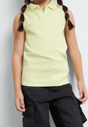 Niño con cabello trenzado que lleva una camisa tipo polo sin mangas de color amarillo pálido y pantalones cargo negros, de pie contra una pared de color claro.