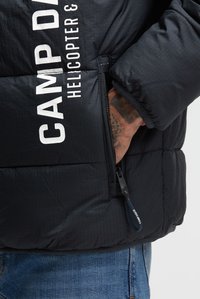 Persoon draagt een zwart gewatteerd jack met de tekst "CAMP DAVID HELICOPTER 3", hand in de zijritszak, blauwe spijkerbroek zichtbaar eronder.