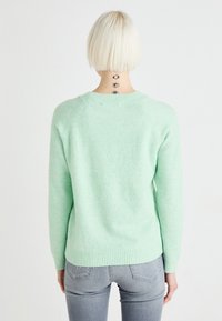 Person med kort blont hår som bär en ljusgrön tröja och grå jeans, visar en vertikal ögon-tatuering på baksidan av sin nacke.