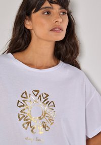 Weißes T-Shirt mit kurzen Ärmeln, das ein großes goldenes Sonnenmotiv und den Text "Immer sonnig" darunter zeigt. Aus weichem, leichtem Stoff.