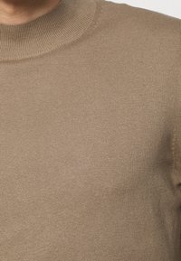 Primo piano di una persona che indossa un maglione a coste beige con colletto alto.