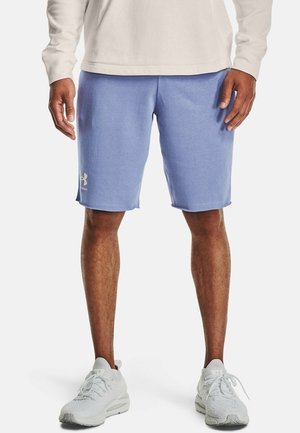 Uomo che indossa pantaloncini sportivi viola chiaro, maglia bianca a maniche lunghe e scarpe da ginnastica bianche, in piedi davanti a uno sfondo neutro.