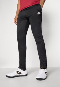 Fekete, slim fit sportnadrág sima anyagból. Fehér logó található a lábon, fehér sportcipővel párosítva.