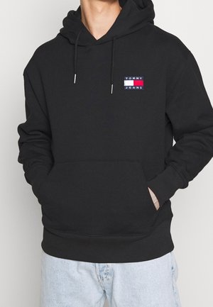 Mann trägt schwarzen Tommy Jeans Hoodie mit Kängurutasche und hellblauen Jeans, Hände in den Taschen, neutraler Hintergrund.