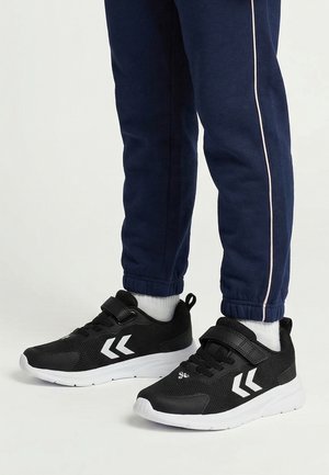 Person iført sorte atletiksneakers med hvide såler og chevron-design, navyblå træningsbukser med sidelænke og hvide sokker.