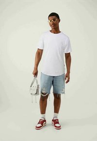 Man i solglasögon, vit t-shirt, slitna denimshorts, röd-vita sneakers, hållande en liten beige crossbody-väska mot en enfärgad bakgrund.