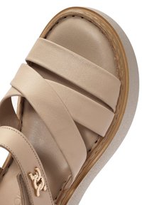 Sanda in pelle beige con ampie fascette incrociate, soletta sagomata e suola piatta di colore chiaro. Accento del logo dorato vicino alla fascetta della caviglia.
