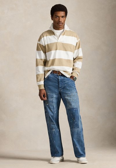 Μπλούζα με μακριά μανίκια σε κρεμ και χακί ρίγες, συνδυασμένη με μπλε τζιν παντελόνια ζωγράφου με πολλαπλές τσέπες και πιτσιλιές μπογιάς. Λευκά αθλητικά παπούτσια.