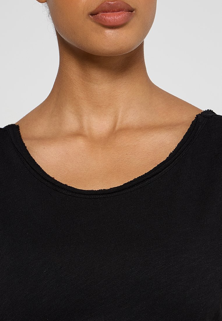 Nahaufnahme des Halses und der Lippen einer Person, die ein schwarzes Rundhalsshirt mit einem rohen Ausschnitt trägt.