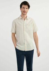 Jasno beżowa koszula z krótkim rękawem, zapinana na guziki, z kieszonką na piersi i gładką teksturą, zestawiona z granatowymi spodniami. Materiał wydaje się lekki.