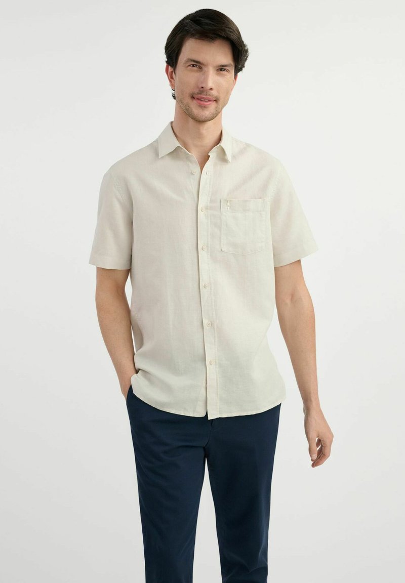 Jasno beżowa koszula z krótkim rękawem, zapinana na guziki, z kieszonką na piersi i gładką teksturą, zestawiona z granatowymi spodniami. Materiał wydaje się lekki.