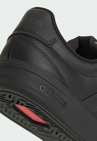 Tacón de zapatilla Adidas negra con cuero texturizado, costuras visibles, logotipo en relieve en la suela y detalle rojo en la banda de rodadura.