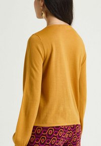 Pull en maille jaune moutarde à manches longues, avec un simple col rond. Porté avec un pantalon à motifs violet et jaune.
