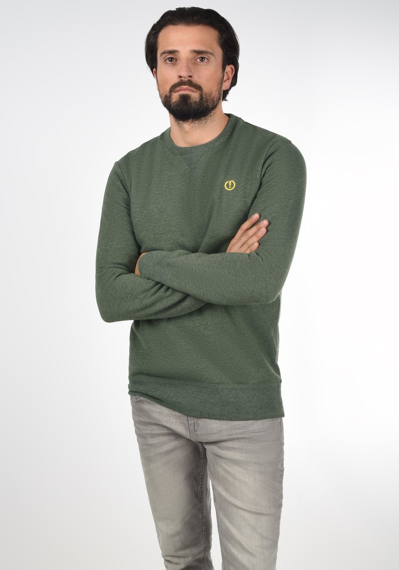 Sweat-shirt vert avec un col rond, tissu texturé, et un petit logo jaune sur la poitrine. Porté avec un jean gris clair.