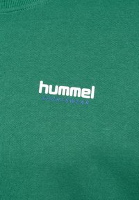 Grön bomullströja med rund halsringning och "hummel"-logotyp i vitt och lila, texturerad tyg med en slät finish.