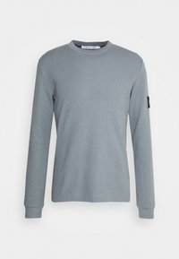 Langarm, hellgrauer Sweatshirt aus strukturiertem Material, runder Halsausschnitt, gerippte Bündchen und ein kleines Logo-Patch am Ärmel.