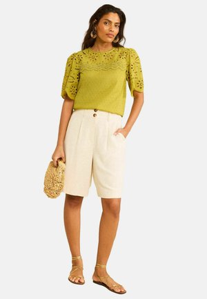 Frau trägt eine grüne Bluse aus Lochspitze, hoch taillierte cremefarbene Shorts mit Knöpfen, hält eine gewebte Tasche und trägt Riemchensandalen.