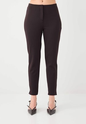 Schwarz geschnittene Slim-Fit-Hose, getragen von einer Person, kombiniert mit schwarzen spitzen High Heels mit Knöchelriemchen, stehend auf einem weißen Boden.