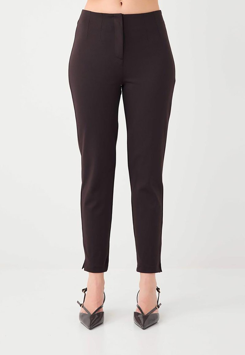 Pantalon noir slim porté par une personne, associé à des talons noirs à bout pointu avec brides à la cheville, debout sur un sol blanc.
