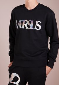 Sudadera negra con un logo holográfico de "VERSUS", cuello redondo y puños de canalé. Acompañada de pantalones negros a juego con detalles similares.