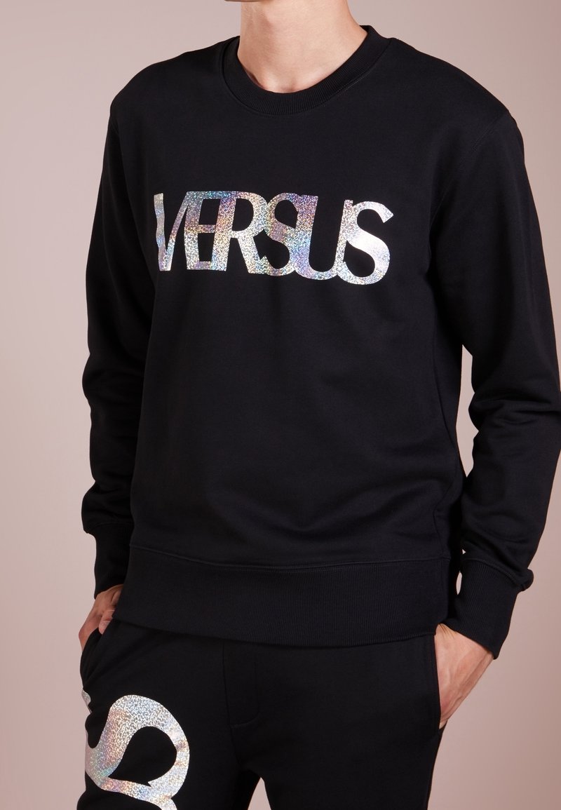 Sudadera negra con un logo holográfico de "VERSUS", cuello redondo y puños de canalé. Acompañada de pantalones negros a juego con detalles similares.