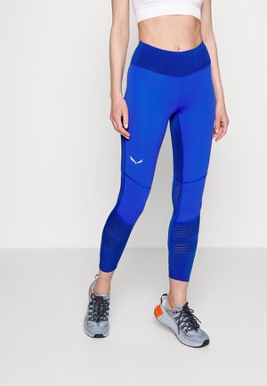 Frau trägt blaue, hoch taillierte Sportleggings und graue Laufschuhe, steht vor einem schlichten weißen Hintergrund.