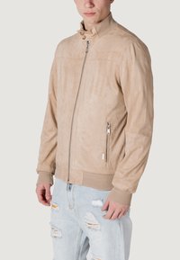 Giacca bomber in suede beige con chiusura a zip frontale, orlo e polsini a costine. Presenta due tasche laterali con zip e un colletto alto.