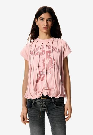 Donna con lunghi capelli scuri indossa una t-shirt rosa con ritagli e grafica e jeans neri sbiaditi con cintura in stile corda, in piedi davanti a uno sfondo bianco.