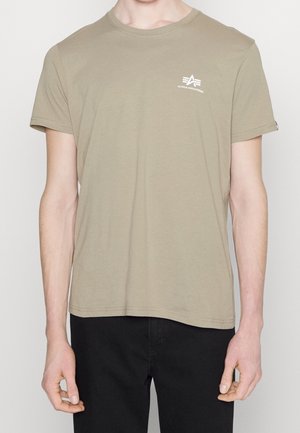 Person trägt ein beiges kurzärmeliges T-Shirt mit einem kleinen weißen Alpha Industries-Logo auf der linken Brust und schwarze Hose.
