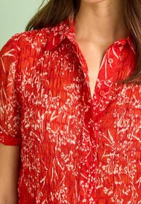 Chemise rouge à manches courtes, avec un design texturé et froissé, arborant un motif floral blanc. Col pointu.