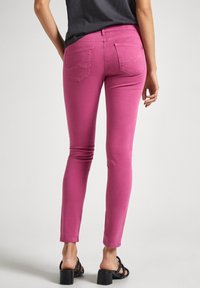 Person som bär magenta slim-fit jeans och svarta klacksandaler, visad från ryggen mot en enfärgad ljus bakgrund.
