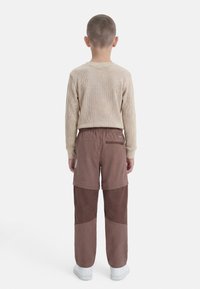 Παιδί που στέκεται με την πλάτη προς τα εμπρός, φορώντας μπεζ μακρυμάνικη μπλούζα, παντελόνια διχρωμίας καφέ και λευκά αθλητικά παπούτσια, μπροστά σε λευκό φόντο.