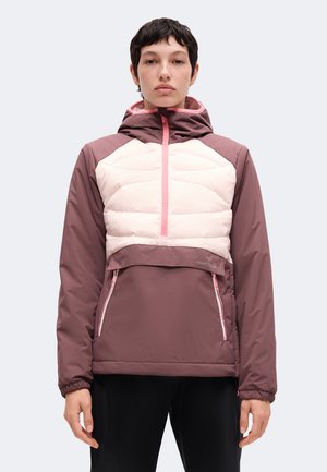 Personne aux cheveux courts et foncés portant une veste à capuche mauve et rose clair avec une fermeture éclair sur le devant et des poches, debout devant un fond uni.
