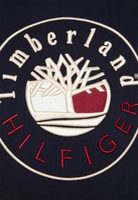 Logo circulaire brodé avec un arbre stylisé blanc et rouge au centre, entouré des textes « Timberland » et « Hilfiger » sur un tissu bleu marine.
