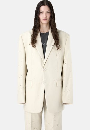 Persona de cabello largo y oscuro que lleva un traje crema oversize con sutiles motas negras y una camiseta gráfica negra contra un fondo liso.