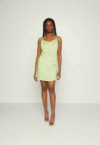 Vestido verde sem mangas com uma forma ajustada, textura suave e detalhe de bolso lateral, estilizado com calçado minimalista.