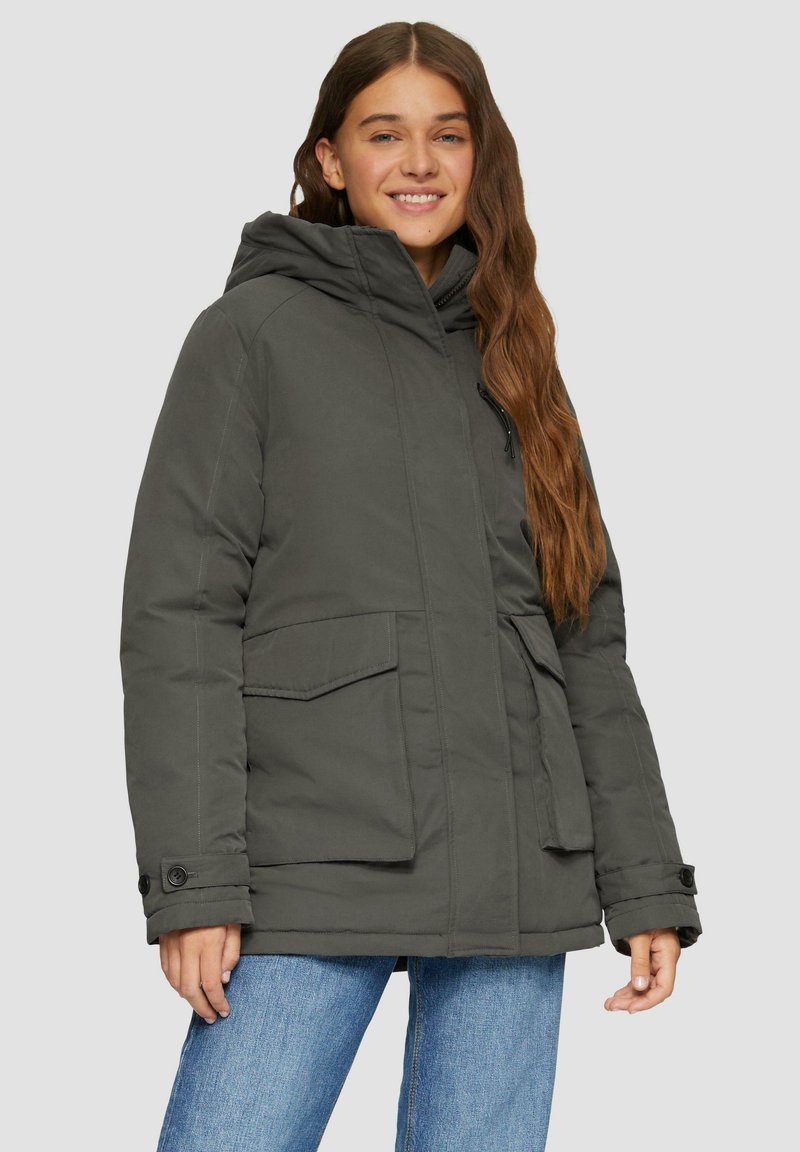 Grijze gewatteerde jas met capuchon, grote voorkruiszakken en knoopmanchetten, gecombineerd met blauwe jeans. Glad stoftextuur.
