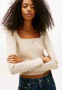Mujer joven con cabello rizado que lleva una blusa de manga larga de canalé beige y jeans de mezclilla oscuros, de pie con los brazos cruzados contra un fondo blanco.