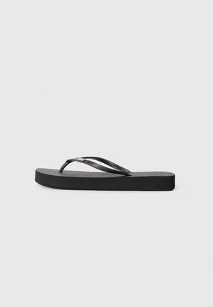 Tongs en caoutchouc noir avec une semelle plate, un lit de pied texturé et deux brides arrondies. Design minimaliste avec un profil élégant.