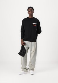 Sort sweatshirt med grafisk logo, lysegrå vidde jeans med kontrasterende syninger, hvide sneakers og en lille sort taske.