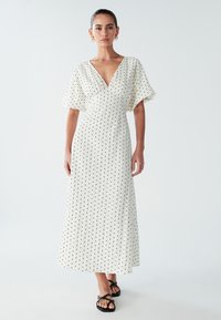 Robe maxi blanche à pois noirs, col en V, manches courtes volantées et tissu texturé ; associée à des sandales noires à brides.